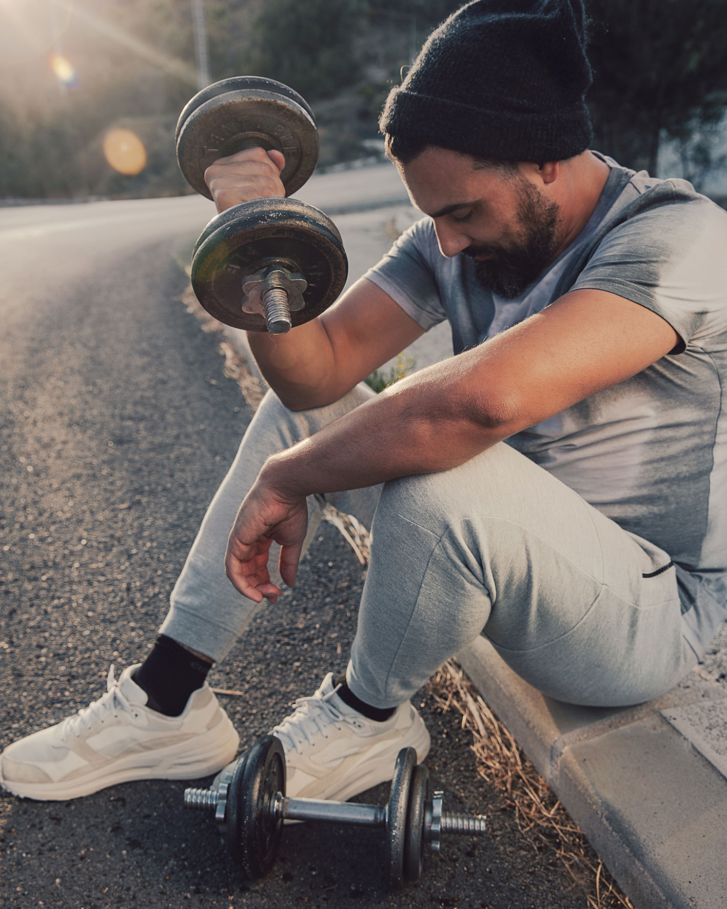 Mann am Straßenrand sitzend mit Hanteln, sportlich gekleidet. Sneaker Allround Farbe schwarz. Elastisch. Gepolsterte Sohle. Ergonomische Anpassung. Schnell trocknend.