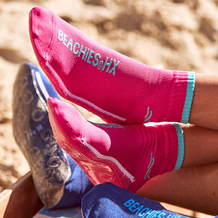 Füße überkreuzt am Strand mit Beachies, den Strand- und Wassersportsocken in Blau und Pink.