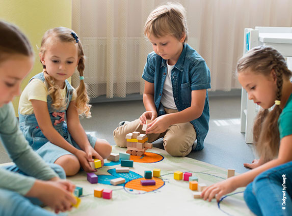 Vier Kinder mit Spielzeug/Holzklötzen auf dem Boden am spielen. Im Hintergrund ein Vorhang und ein Kindertisch.