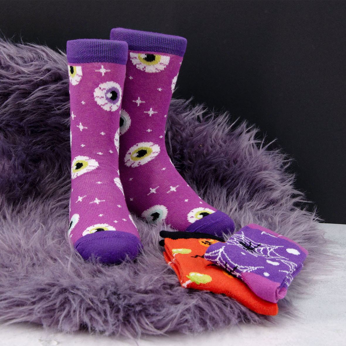 Halloween Herbst Socken lila mit Augen/Augäpfeln und Sternen auf einem lilafarbigen Teppich auf einer weißen Platte neben Socken in orange und lila mit den Motiven Kürbis und Lollis und Spinnen.