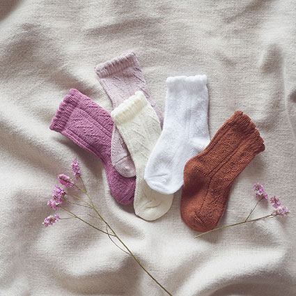 Kindersocken in Traforatomuster in Farben Beere, Beige, Latte, Weiß, und Camel, auf einer cremefarbenden Leinendecke mit Blumen dekoriert.