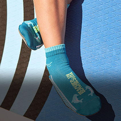 Kind sitzend am Strand auf dem Surfbrett mit den Beachies, der Strand- und Wassersportsocke in Lapis-Blau mit Delphin.