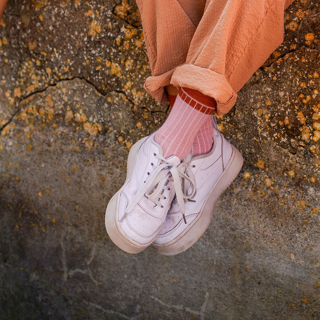 Kind mit überkreuzten Beinen und weißen Turnschuhen auf Mauer sitzend. Farben Rosa, Orange.