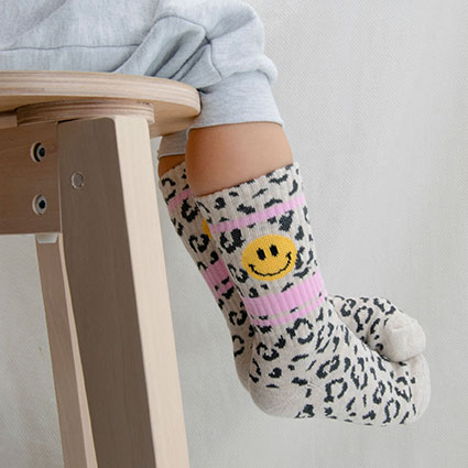 Kinderbeine sitzend auf einem Stuhl mit überschlagenen Fußspitzen und den Leo Smilie, Mini Me Tennis Socken.